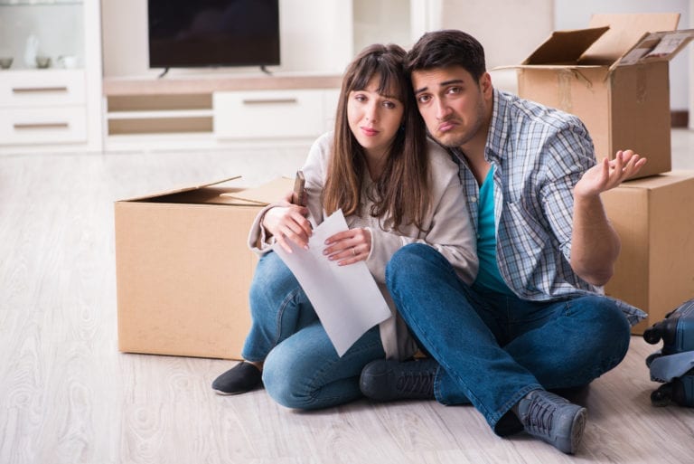A man and a woman sit on the floor of an apartment with moving boxes open in the background while making sad faces. The woman is holding an illegible letter, which is presumably the cause of their sad faces.